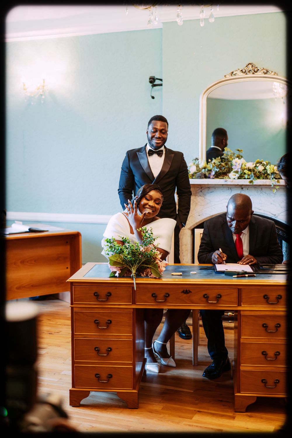 Joyful wedding ceremony moment at a vintage wooden desk with ornate mirror and blue walls in elegant venue.