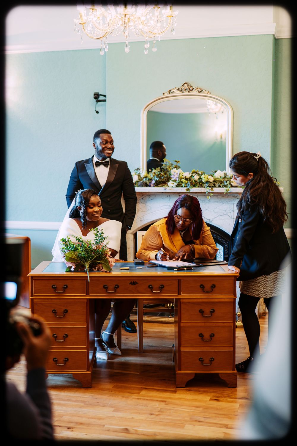 Couple signing wedding documents at wooden desk with floral arrangements in elegant room with blue walls.