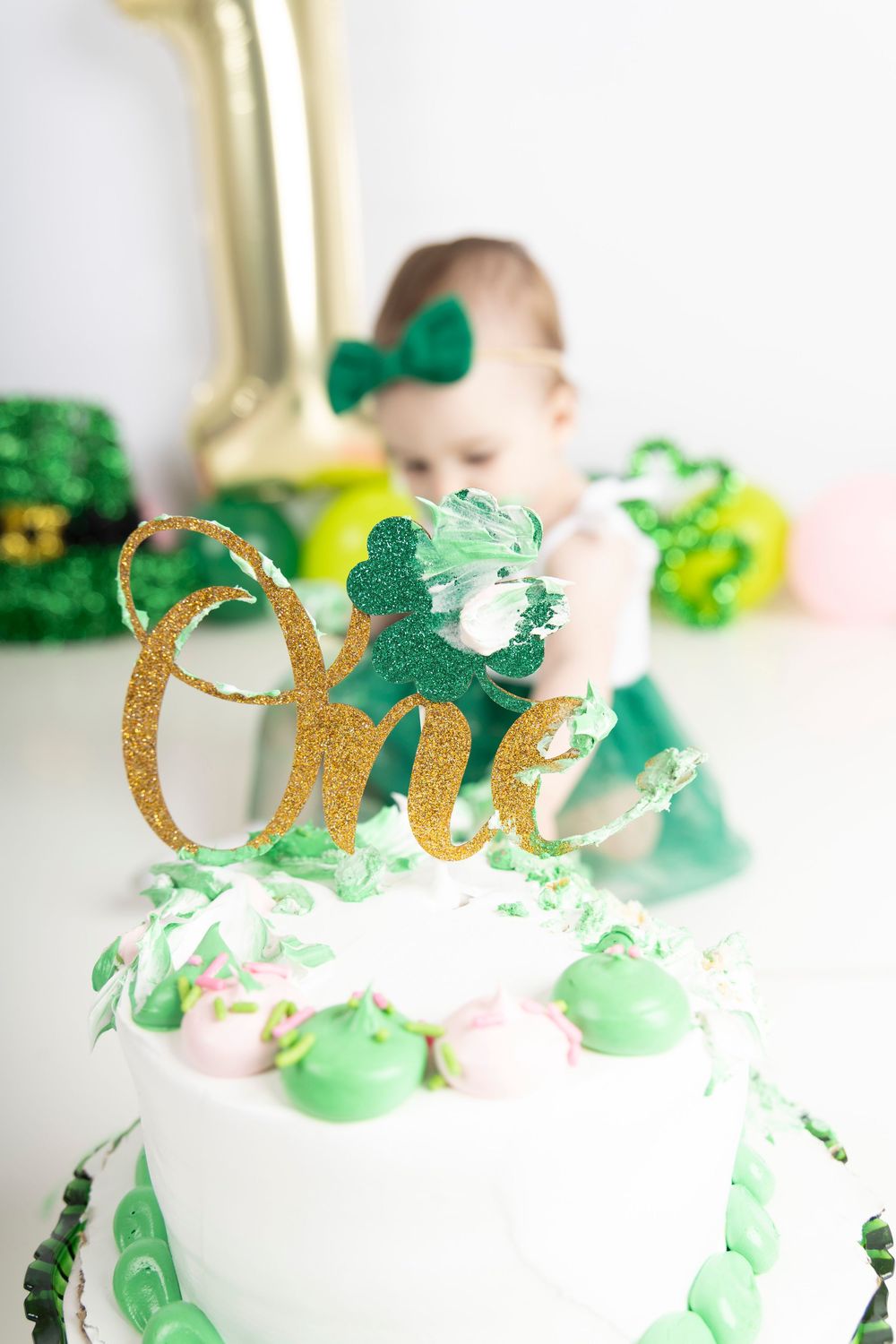 White cake with green decorations and gold 'One' topper for St. Patrick's Day themed first birthday celebration.