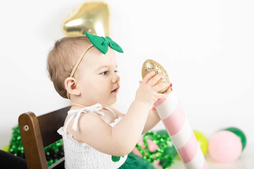 Baby in white dress with green bow celebrates St Patrick's Day with festive decorations and props.