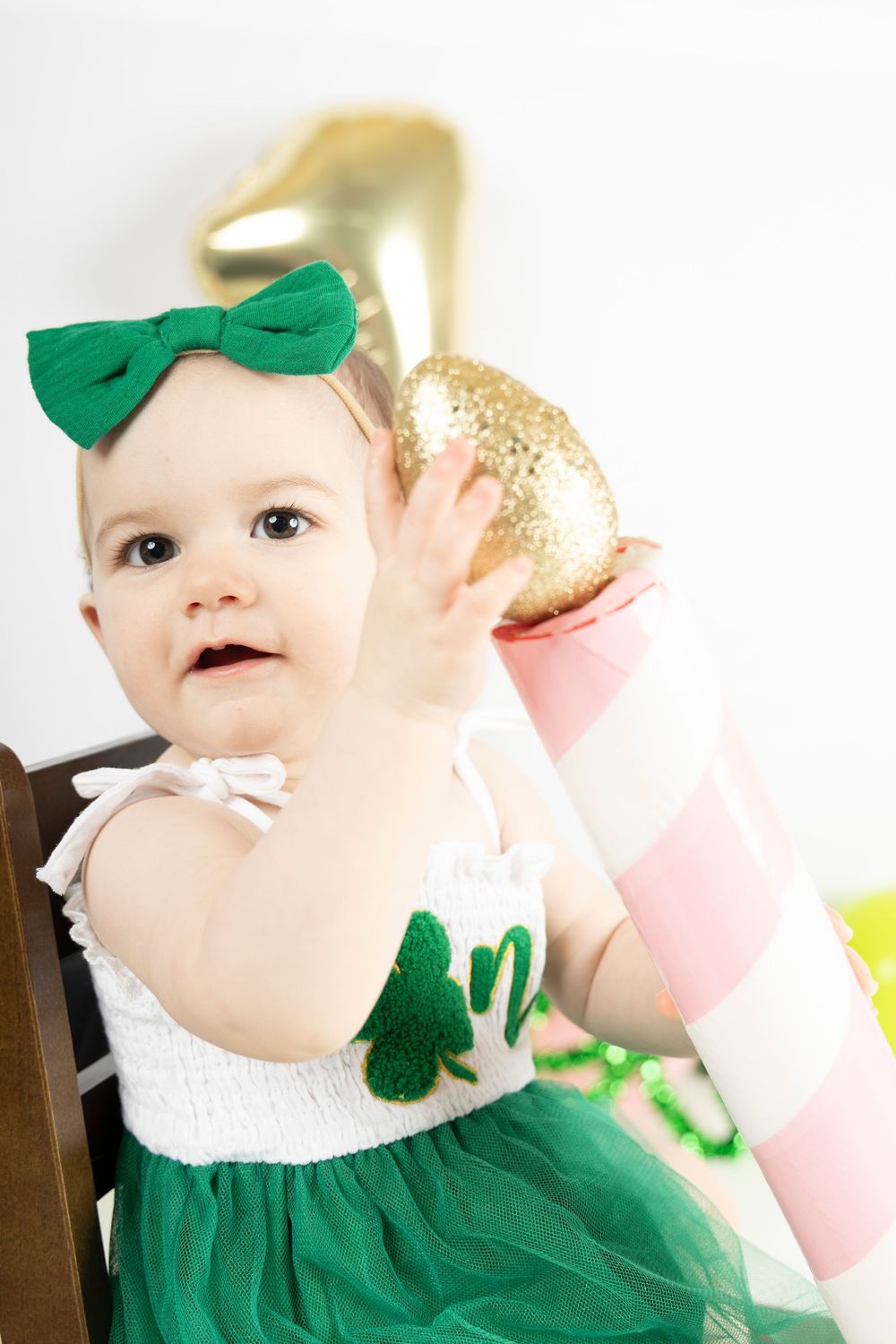 A baby in a green and white St. Patrick's Day outfit reaches for a large gold number one balloon.