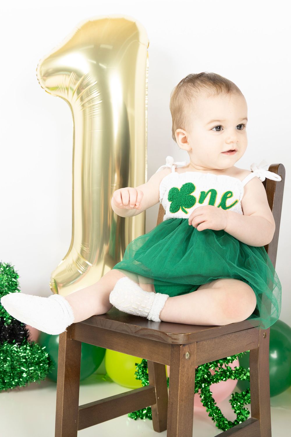 Baby in green outfit sits on wooden chair with gold number one balloon and St. Patrick's Day decorations.