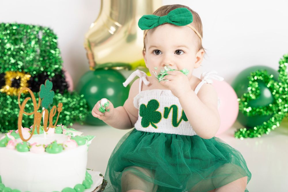 A baby in festive green attire sits near a gold number one balloon and St. Patrick's Day decorations.