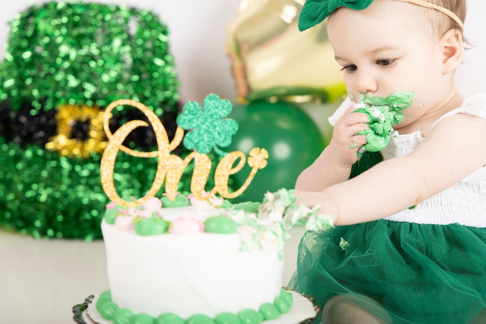 A baby in green dress reaches for a white birthday cake decorated with gold 'One' topper and shamrocks.