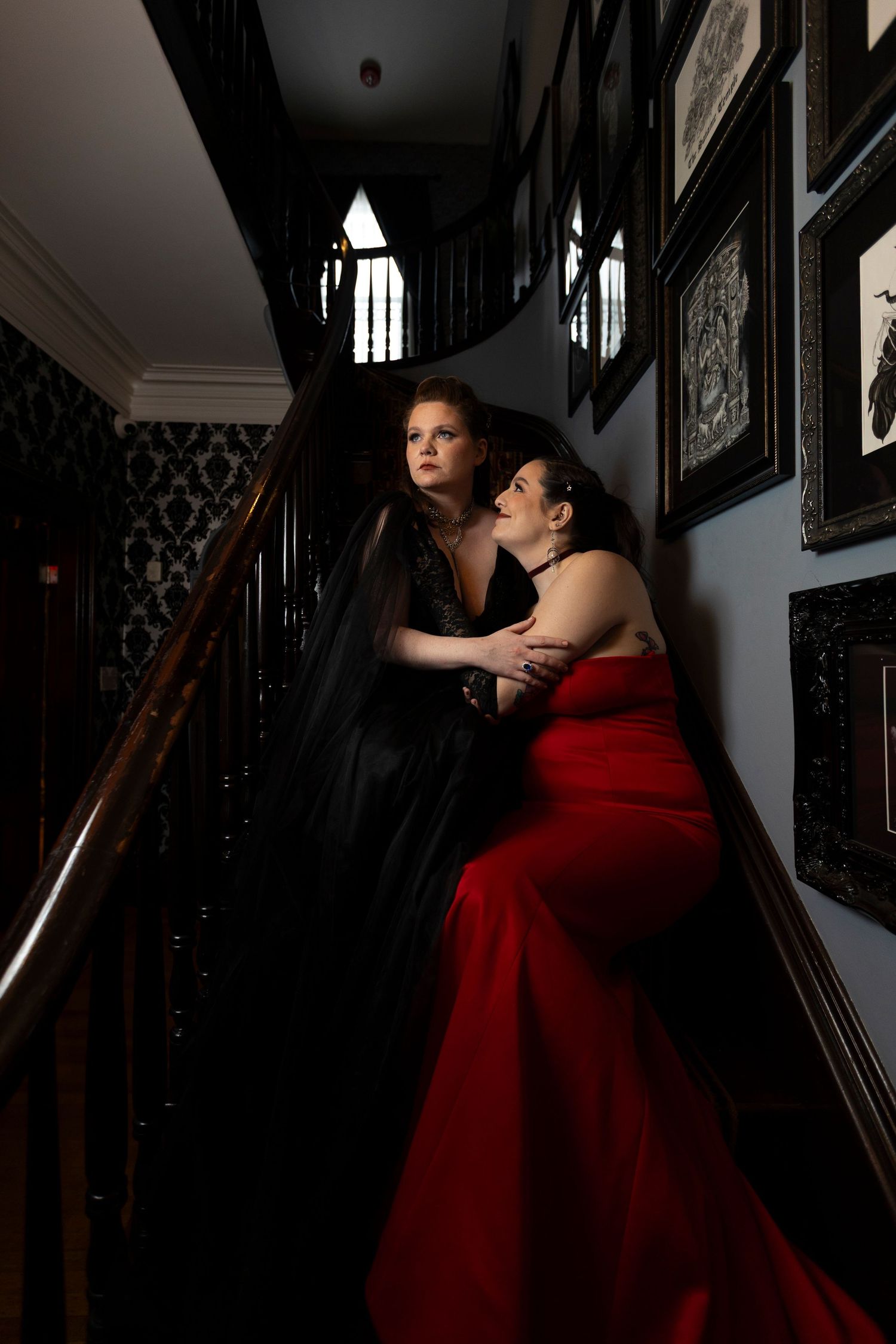 A woman in a flowing red gown poses dramatically on a dark staircase with ornate railings in moody lighting.