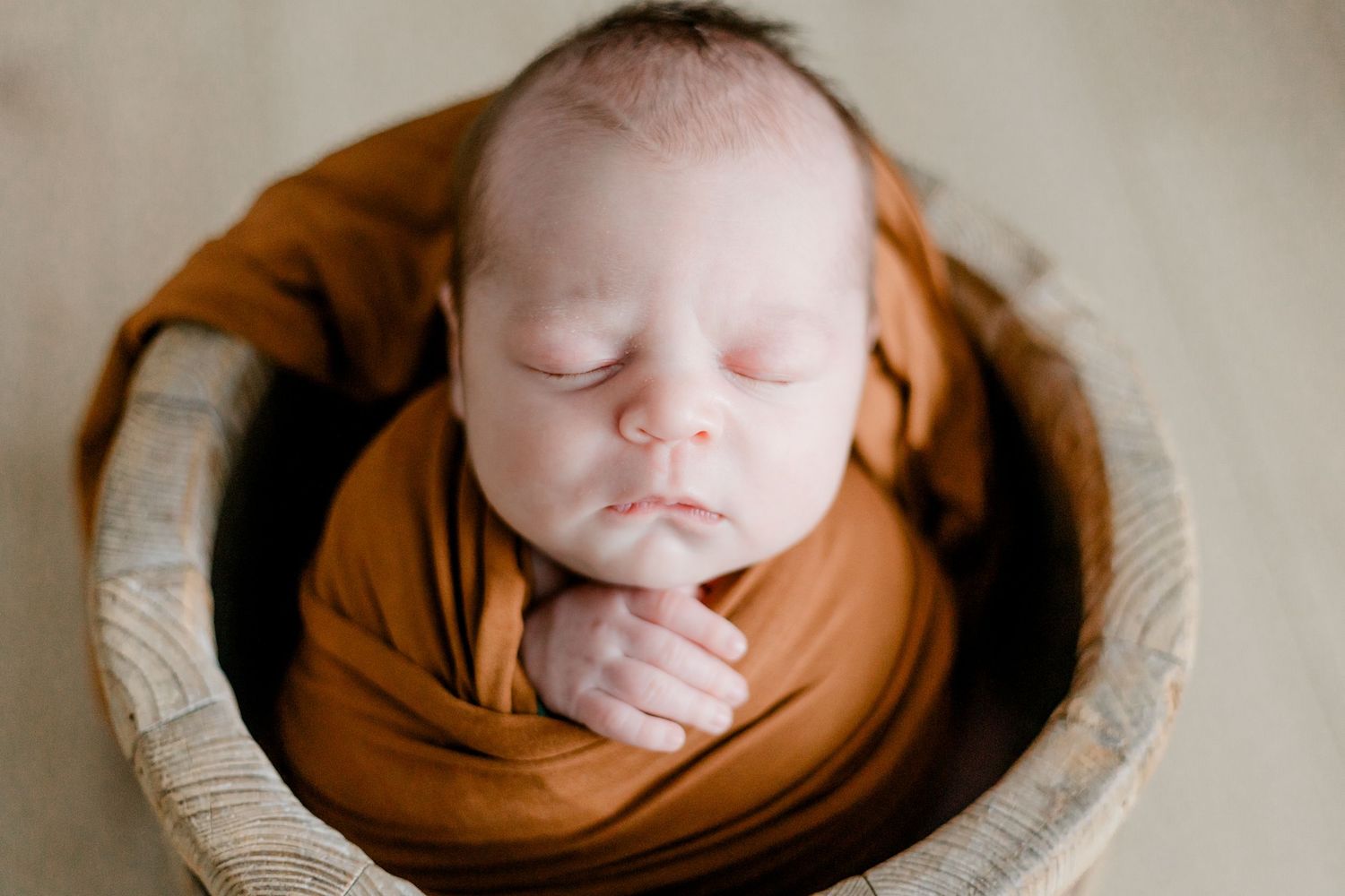 Sleeping newborn baby swaddled in burnt orange wrap, nestled in a woven basket for a professional photo.