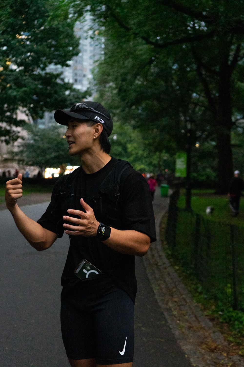 Runner in black athletic wear gestures while exercising on a tree-lined path in a park.