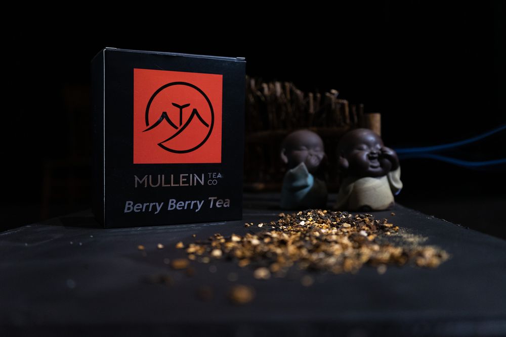 A box of Nullein Berry Berry Tea with loose tea leaves scattered on a dark surface beside decorative elements.