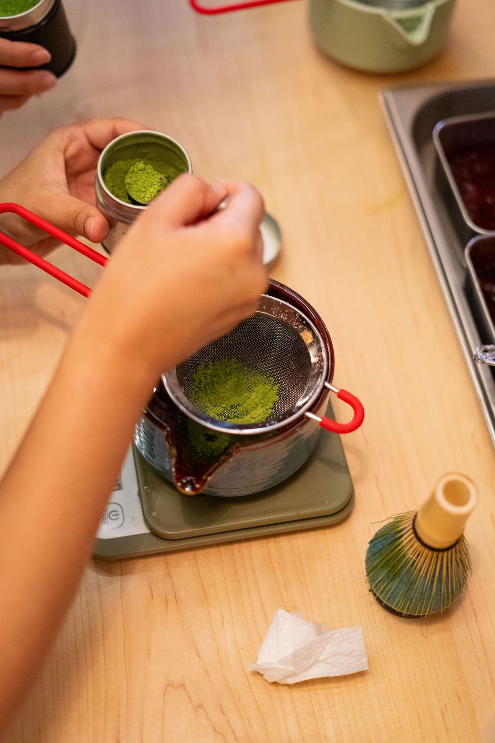Hands preparing traditional matcha green tea with bamboo whisk and strainer on wooden table with digital scale.