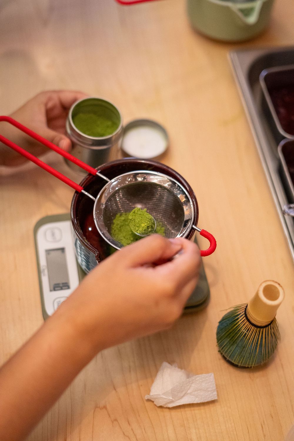 Hands preparing matcha green tea powder in a bowl with a bamboo whisk on a wooden surface.