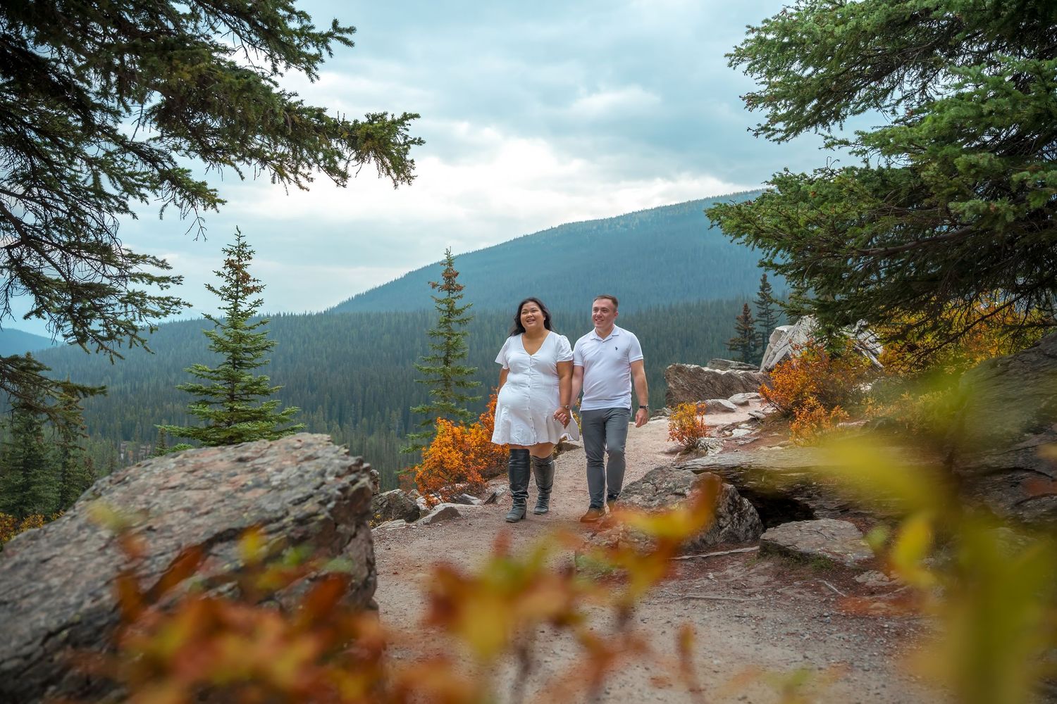 A couple walks along a scenic mountain overlook trail with evergreen trees and autumn foliage in the foreground.