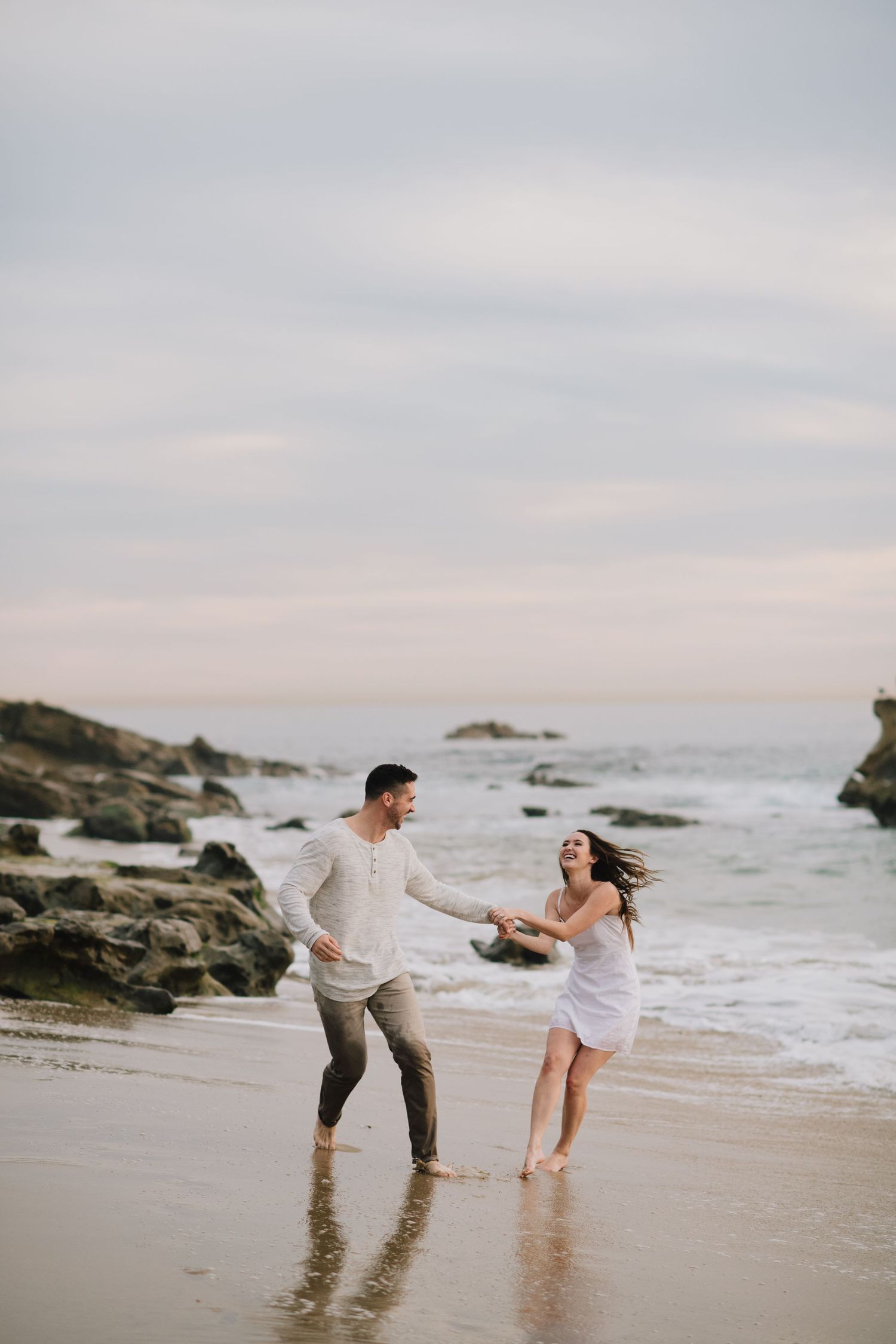 Joyful couple holds hands and dances barefoot on a rocky beach at sunset.