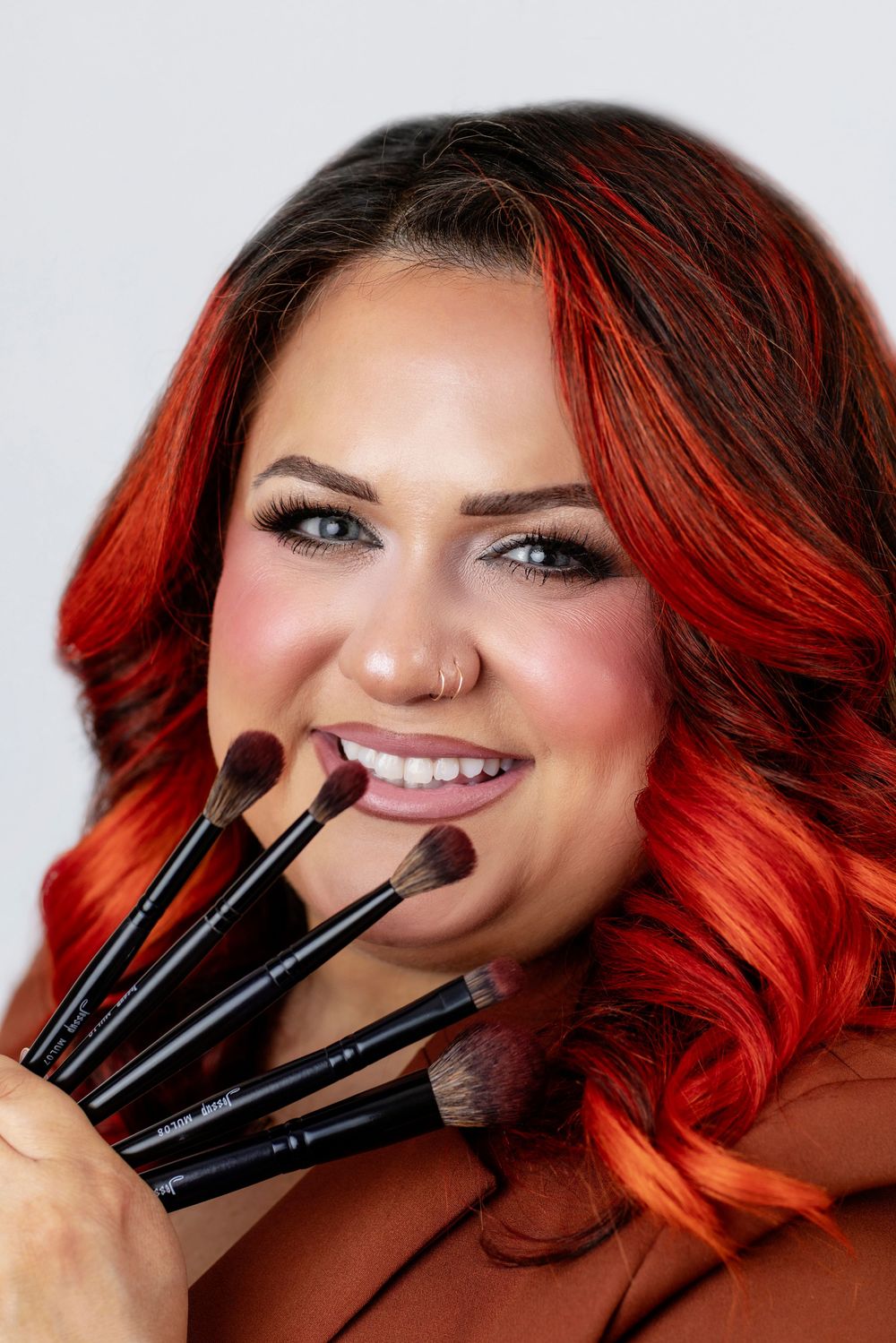 Makeup artist holding three black brushes against vibrant red hair in a creative beauty portrait series.