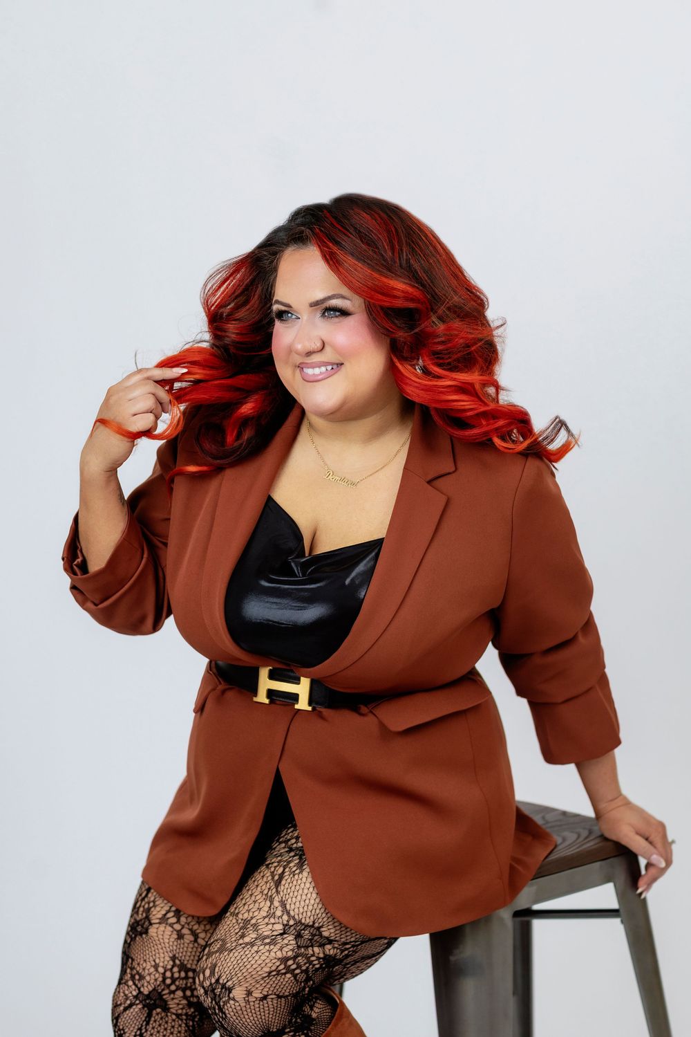 A person with vibrant red hair wearing a brown blazer and lace tights poses confidently on a stool against a white backdrop.