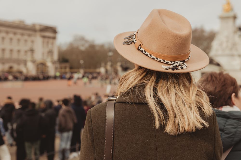 A person wearing a brown fedora hat with leopard print band watches a crowded public square from behind.