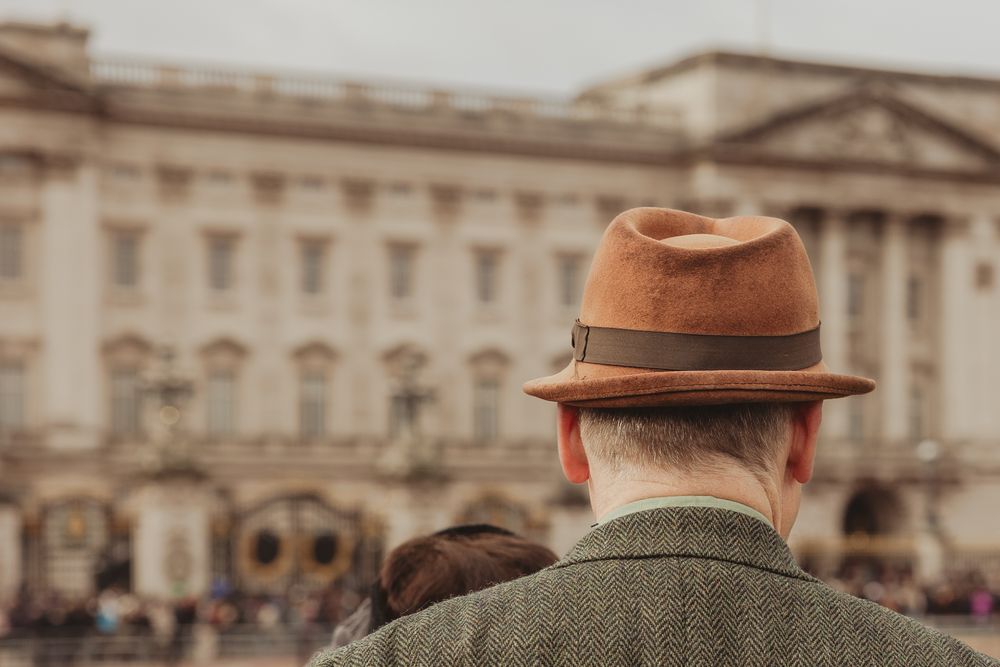 A person wearing a brown fedora hat views Buckingham Palace from behind in London, England.