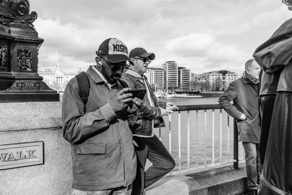 People in caps check their phones while standing along a waterfront railing with a city skyline in the background.