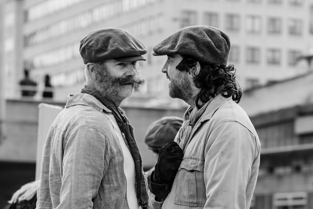 Two men in vintage clothing and flat caps stand close together in a black and white photograph.