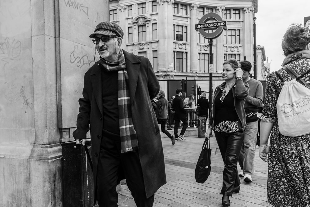 Black and white street photography shows people walking past a building facade with arched columns and doorways.