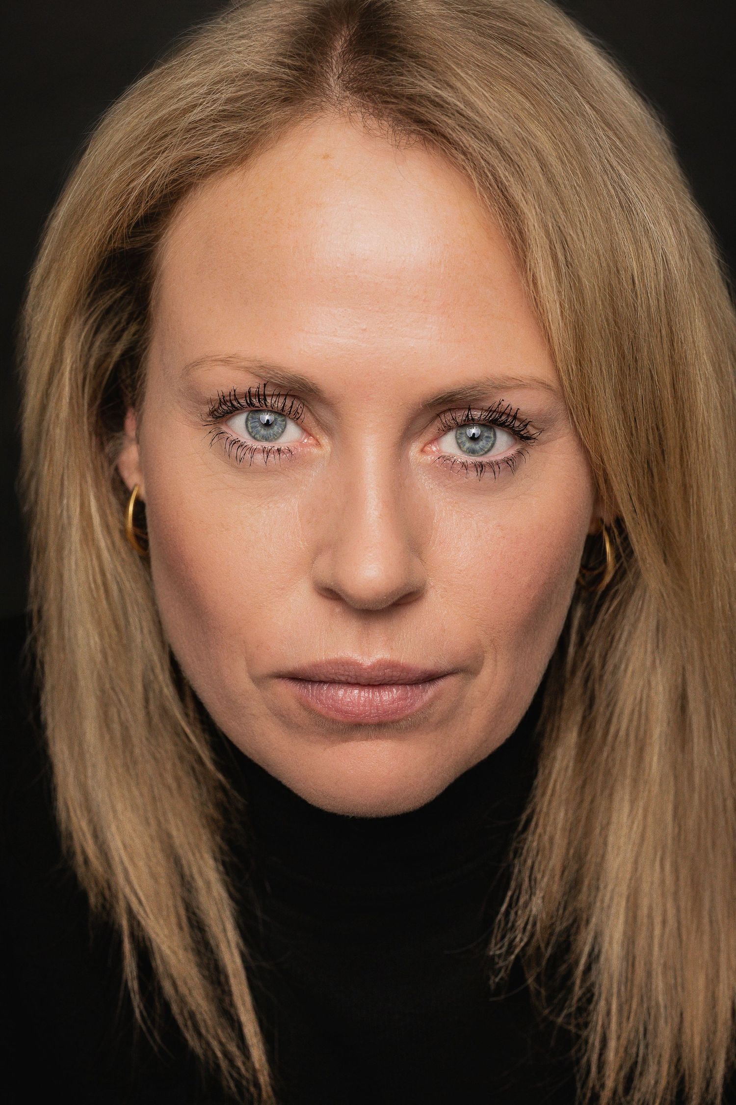 Professional headshot portrait with blonde hair and striking blue eyes against dark background.