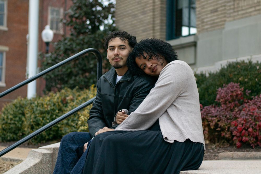 Two people embrace on concrete steps near landscaped buildings.