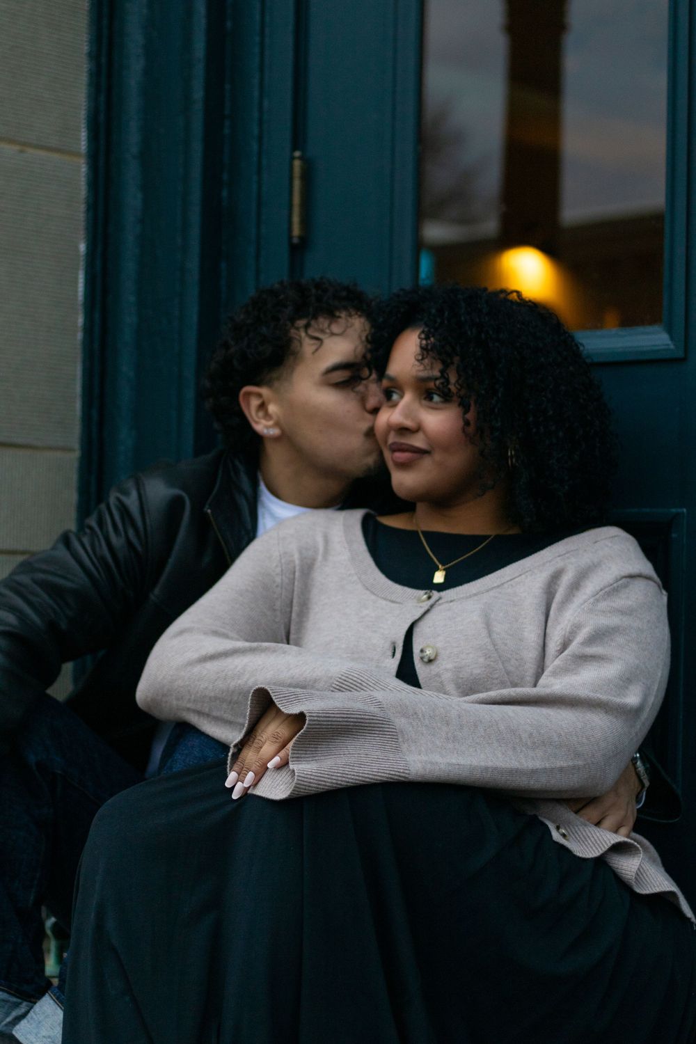 A couple shares a tender moment in front of illuminated storefront windows at dusk.
