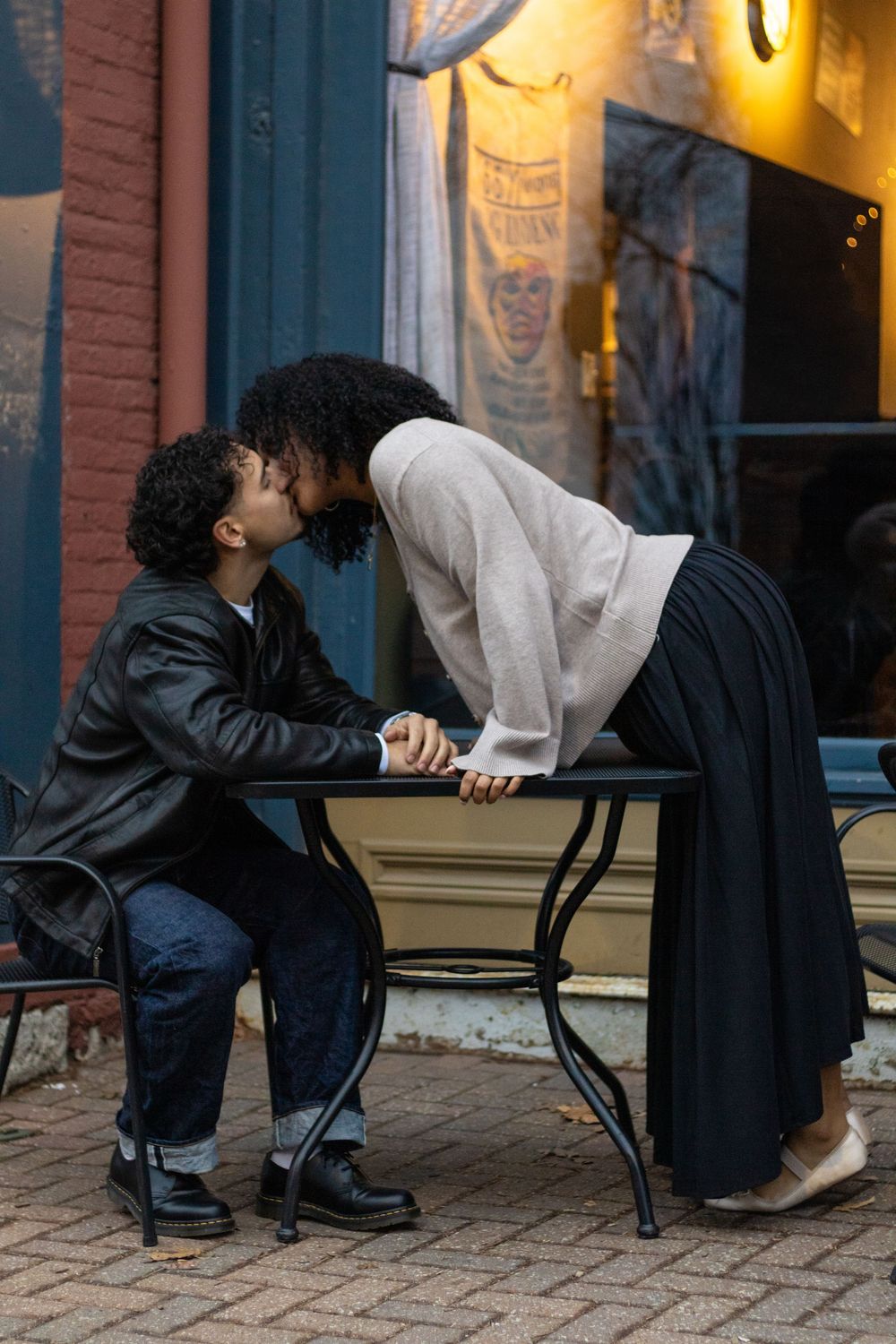 Two people share an intimate conversation on a bench outside a brick building with warm lighting.