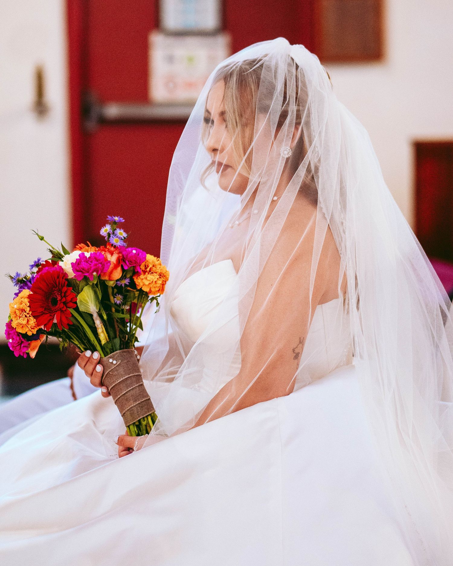 A bride in a white wedding veil holds a vibrant bouquet of red, pink, and orange flowers against a light background.