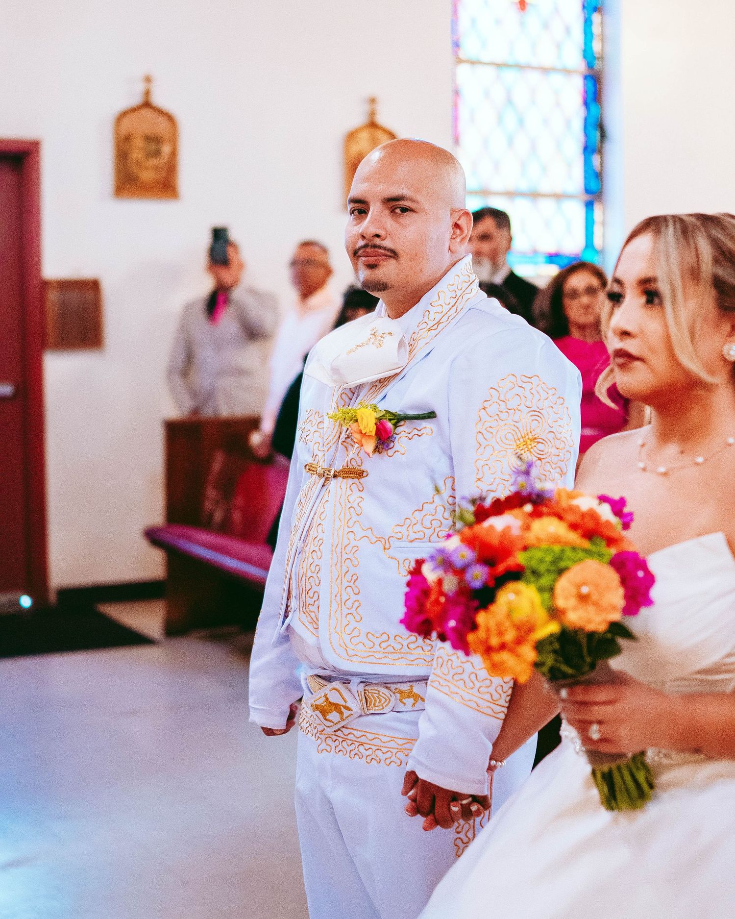 A couple in white and gold wedding attire standing together in a bright church interior with colorful floral bouquet.
