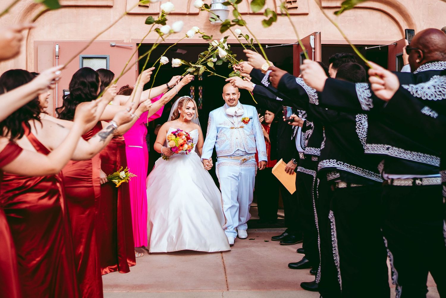 Wedding party poses with bridesmaids in red dresses and groomsmen in dark suits at an outdoor venue.