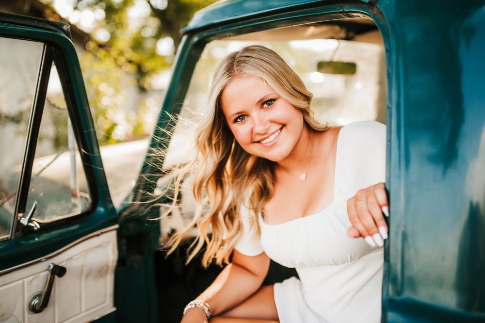 Woman in white dress smiling from vintage turquoise truck during photo session.