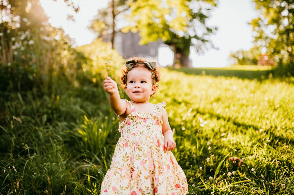 Young child in floral dress holding dandelion in sunlit garden.
