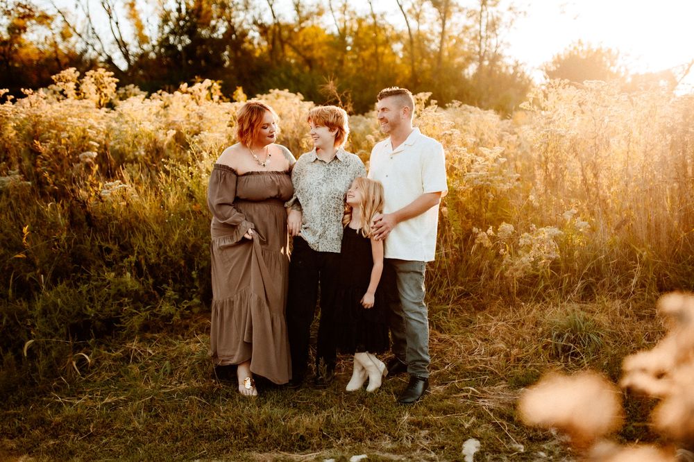 A group of people walk through a sunlit meadow at golden hour with tall grass and trees in the background.