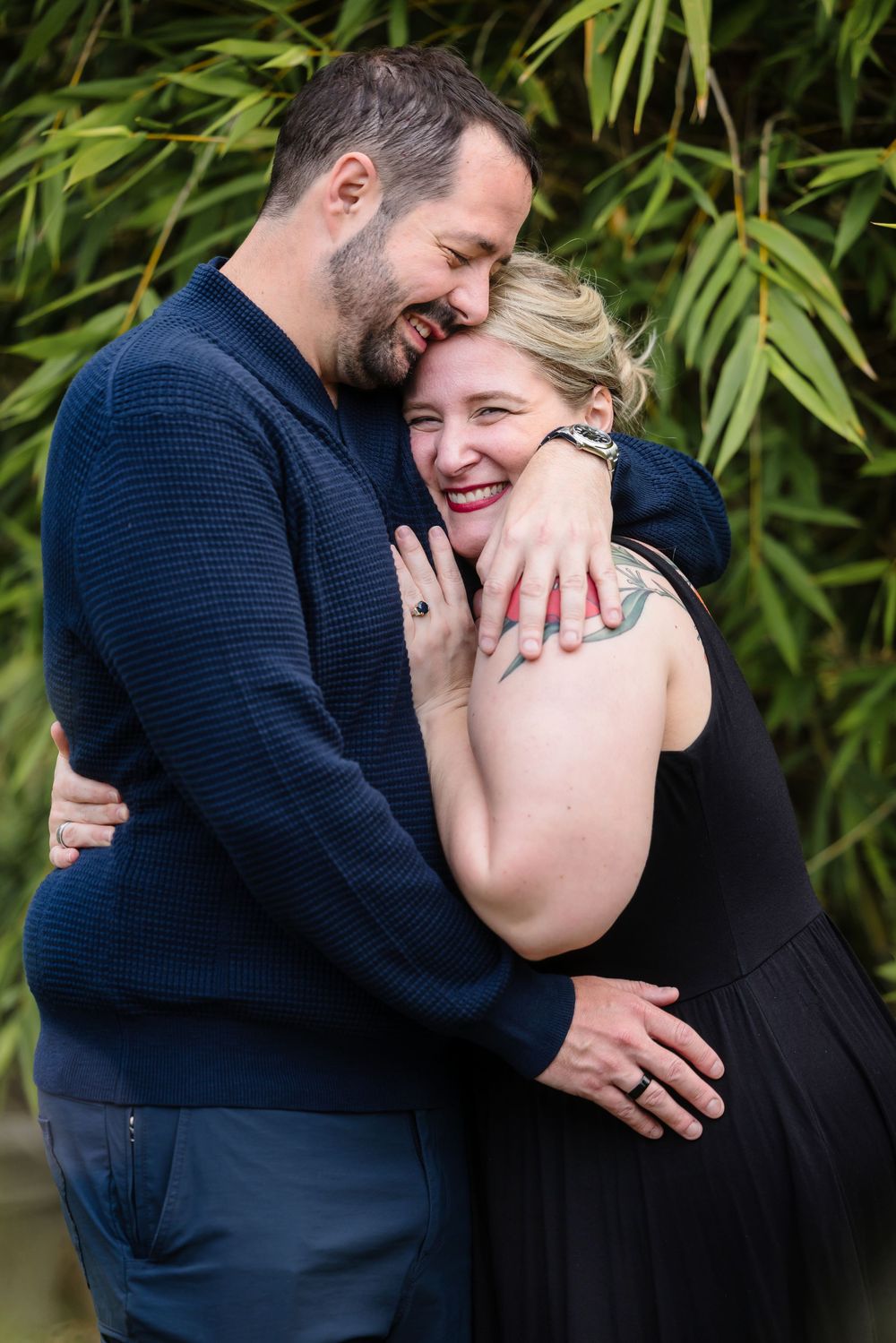 A couple in dark clothing shares intimate laughs against a backdrop of green bamboo leaves.