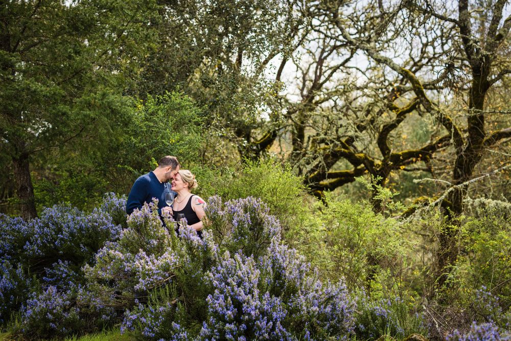 Couple embraces in a romantic outdoor garden setting surrounded by blooming purple flowers and lush greenery.