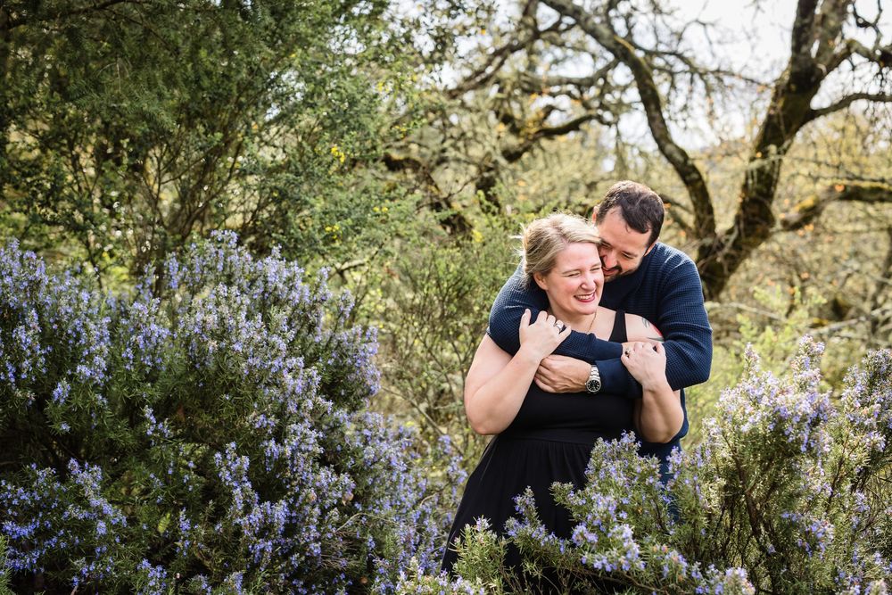 A couple embraces in a romantic moment surrounded by blooming spring flowers and trees in a natural outdoor setting.