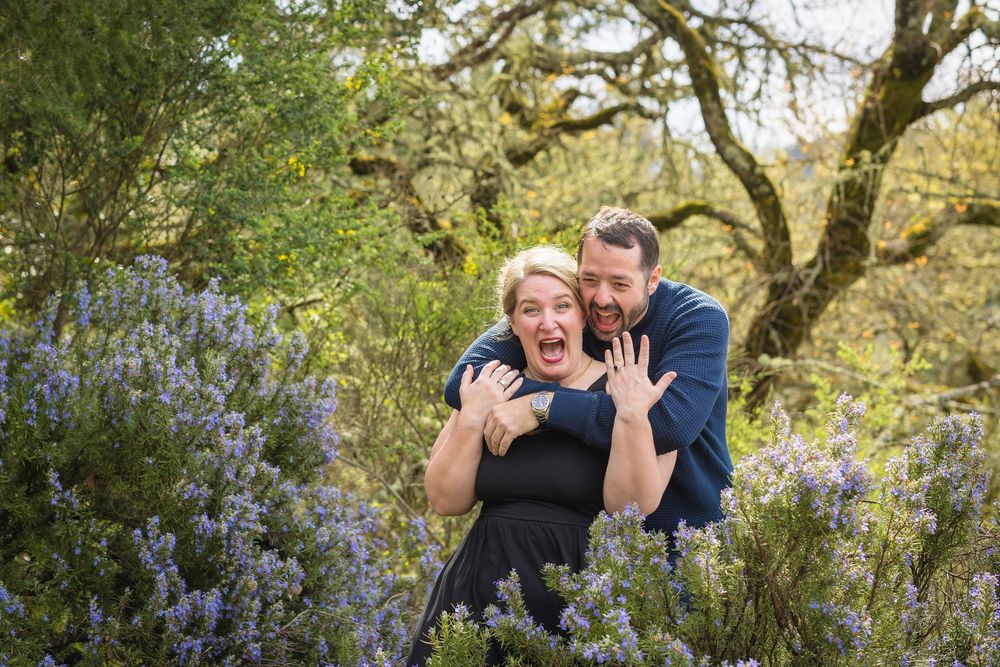 A couple shares joyful laughter together in a lush garden setting with blooming bluebells and oak trees.