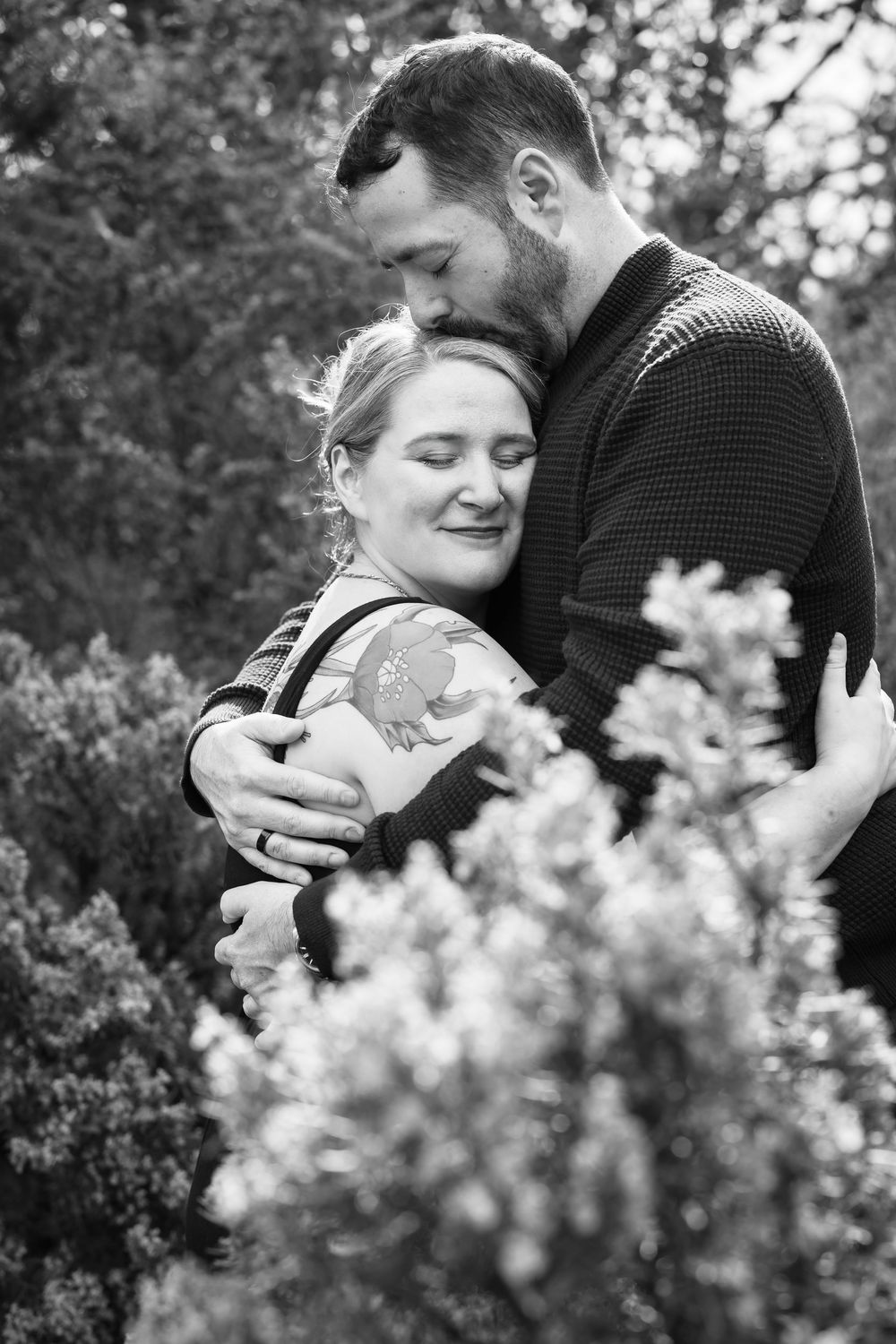 A romantic couple shares tender moments among blooming white flowers in a black and white photograph.
