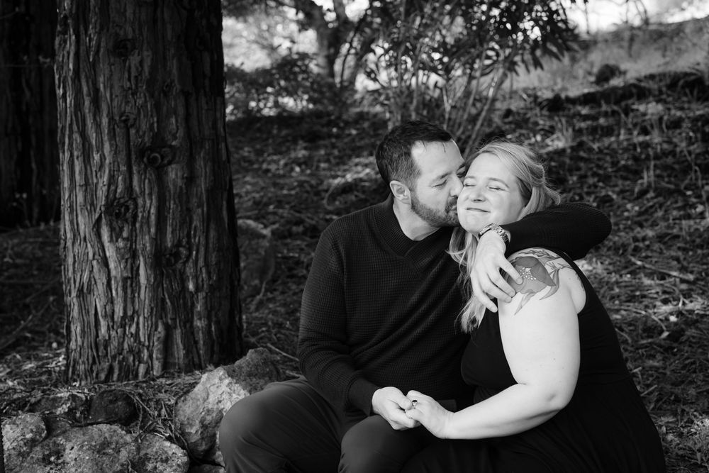 A romantic black and white portrait of a couple sharing an intimate moment while sitting outdoors near a tree.