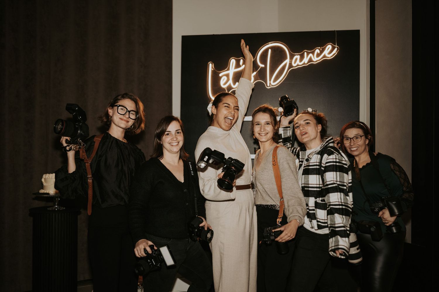 A group of people gather at a dance event with a neon sign visible in the dimly lit venue.