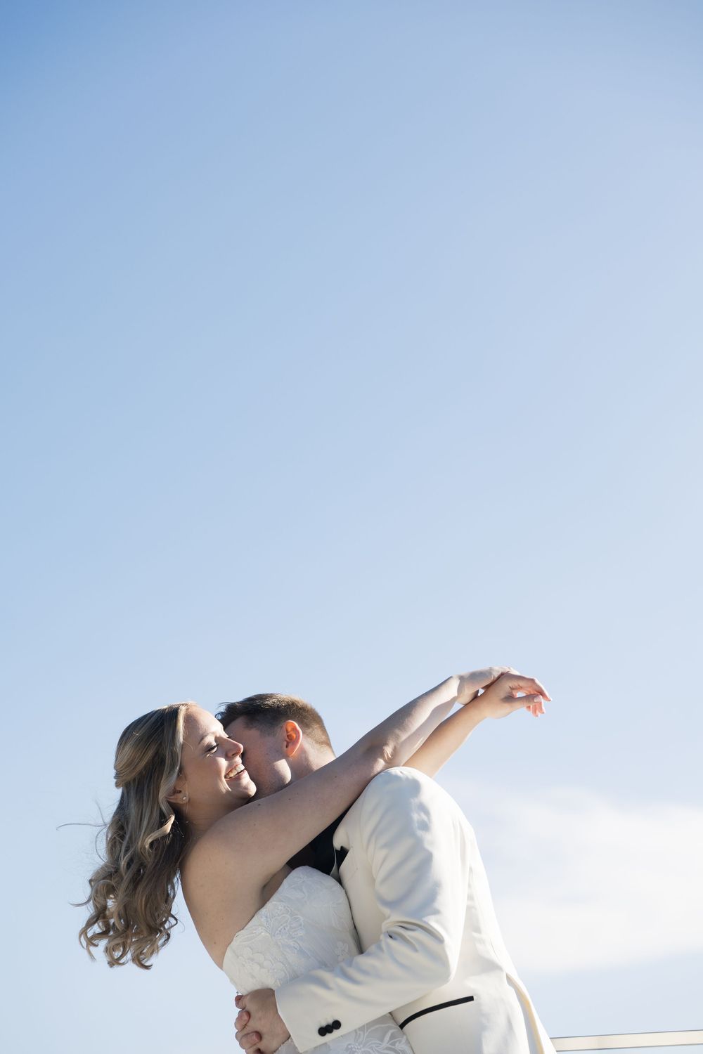 A romantic couple in formal attire dance together against a bright blue sky background.