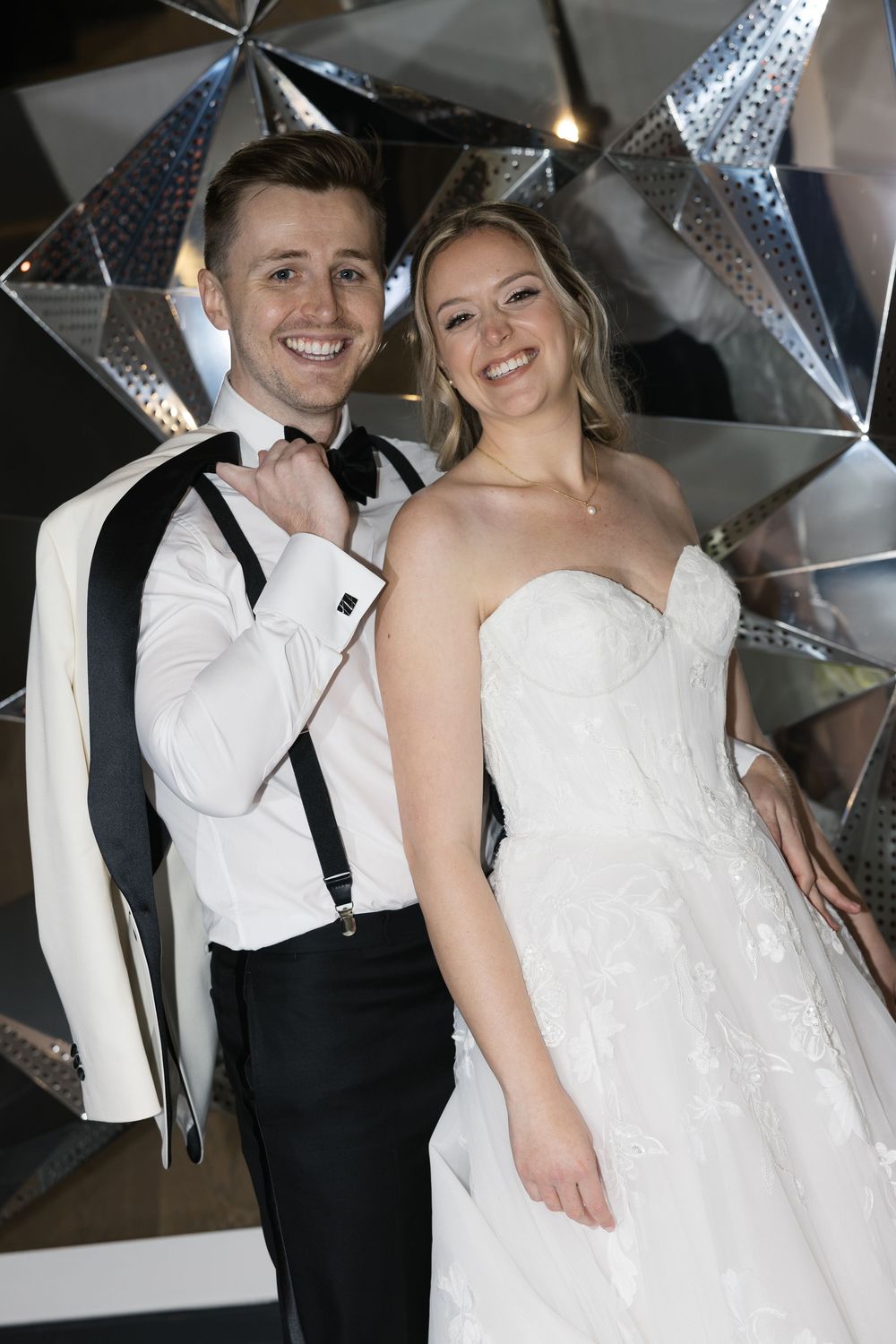 A bride and groom share intimate wedding photos in front of a metallic backdrop with dramatic lighting.