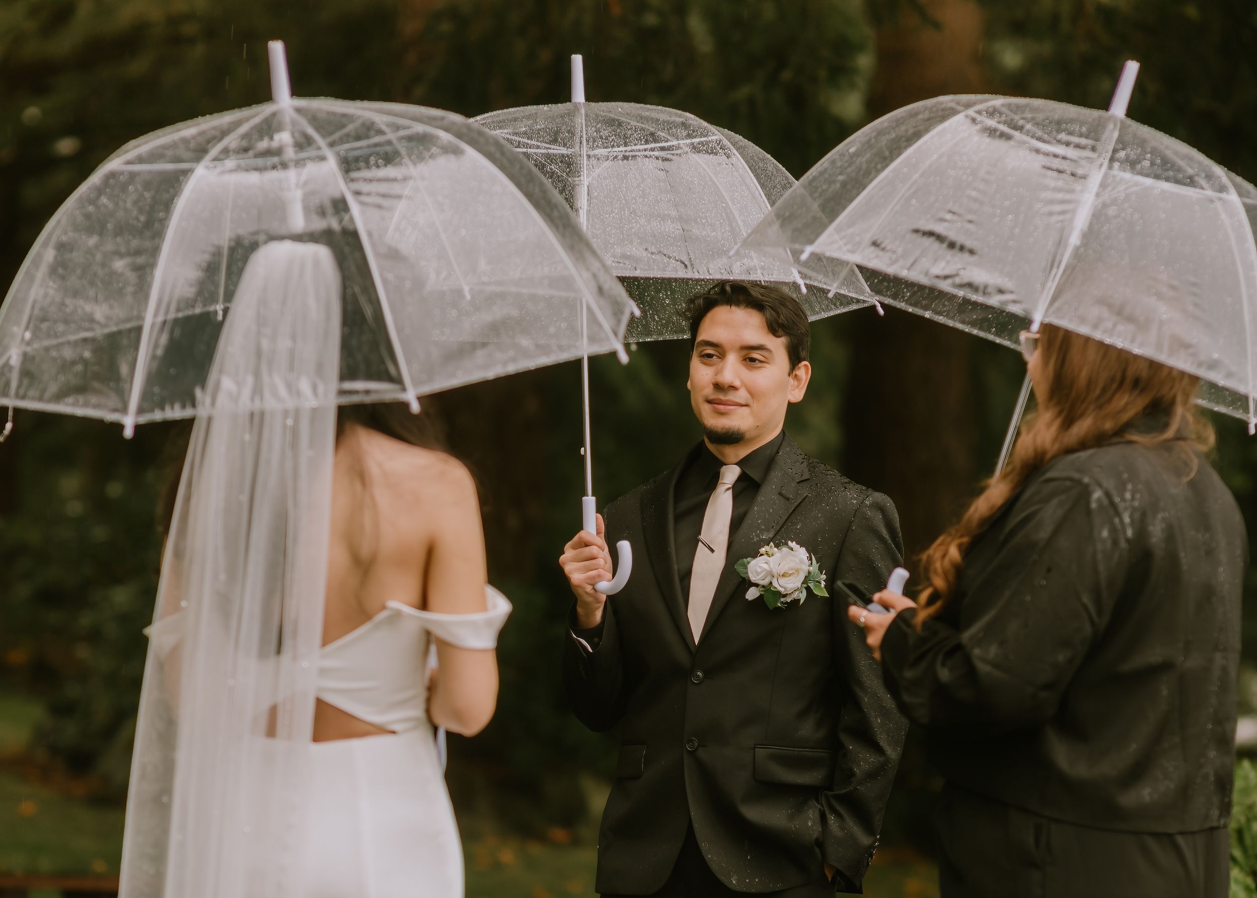 Wedding party members hold clear umbrellas while standing together in the rain during an outdoor ceremony.