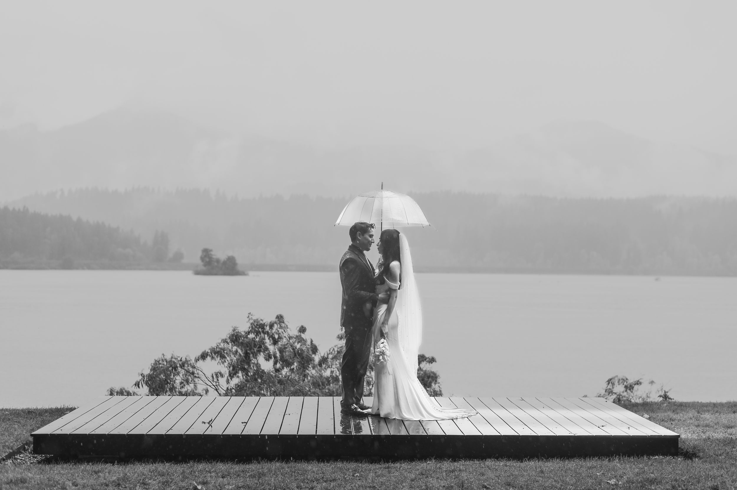 A couple stands together under a white umbrella on a dock overlooking a misty lake in black and white.