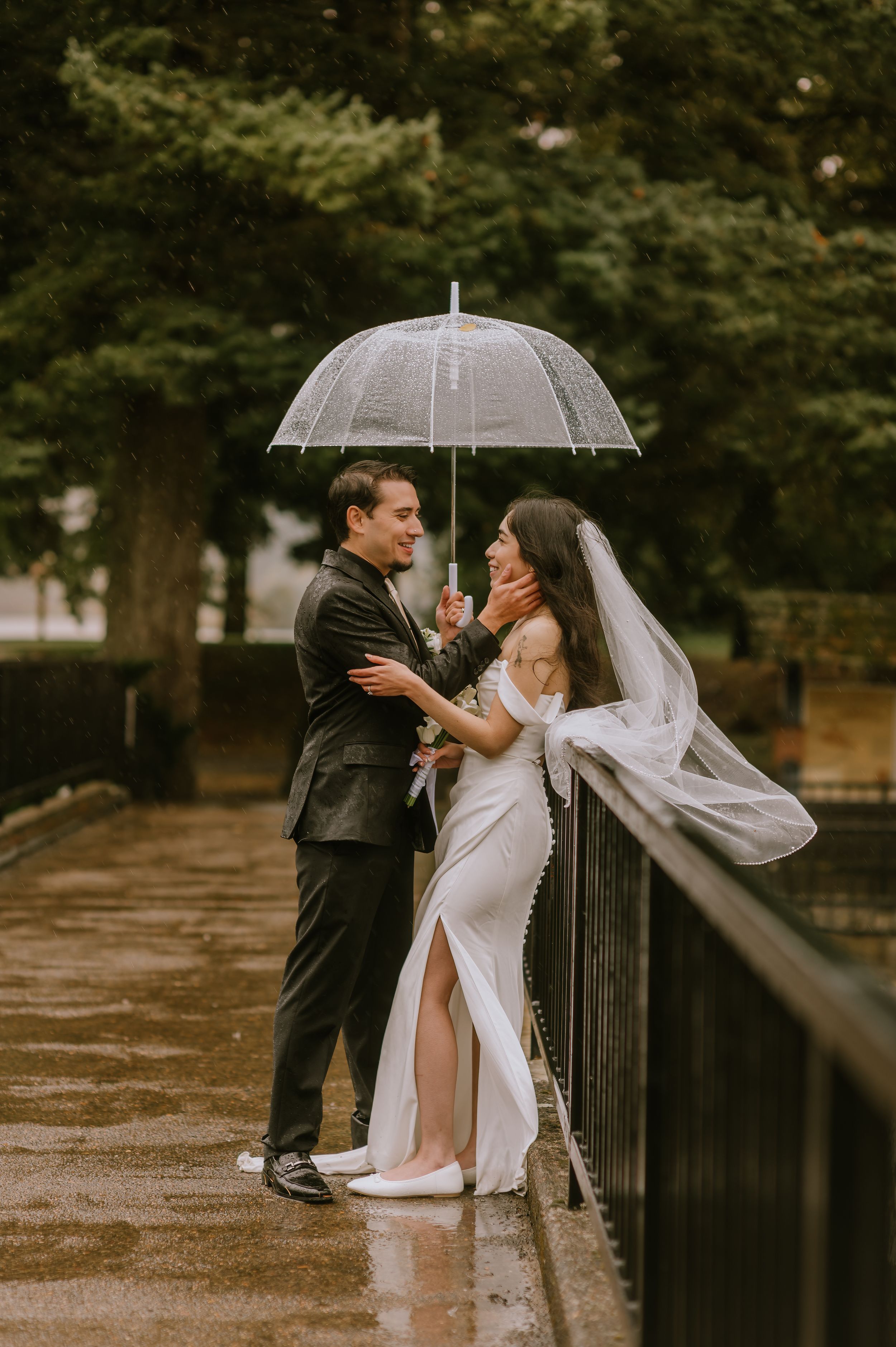 A couple shares an intimate moment under a clear umbrella on a rainy day along a metal railing.