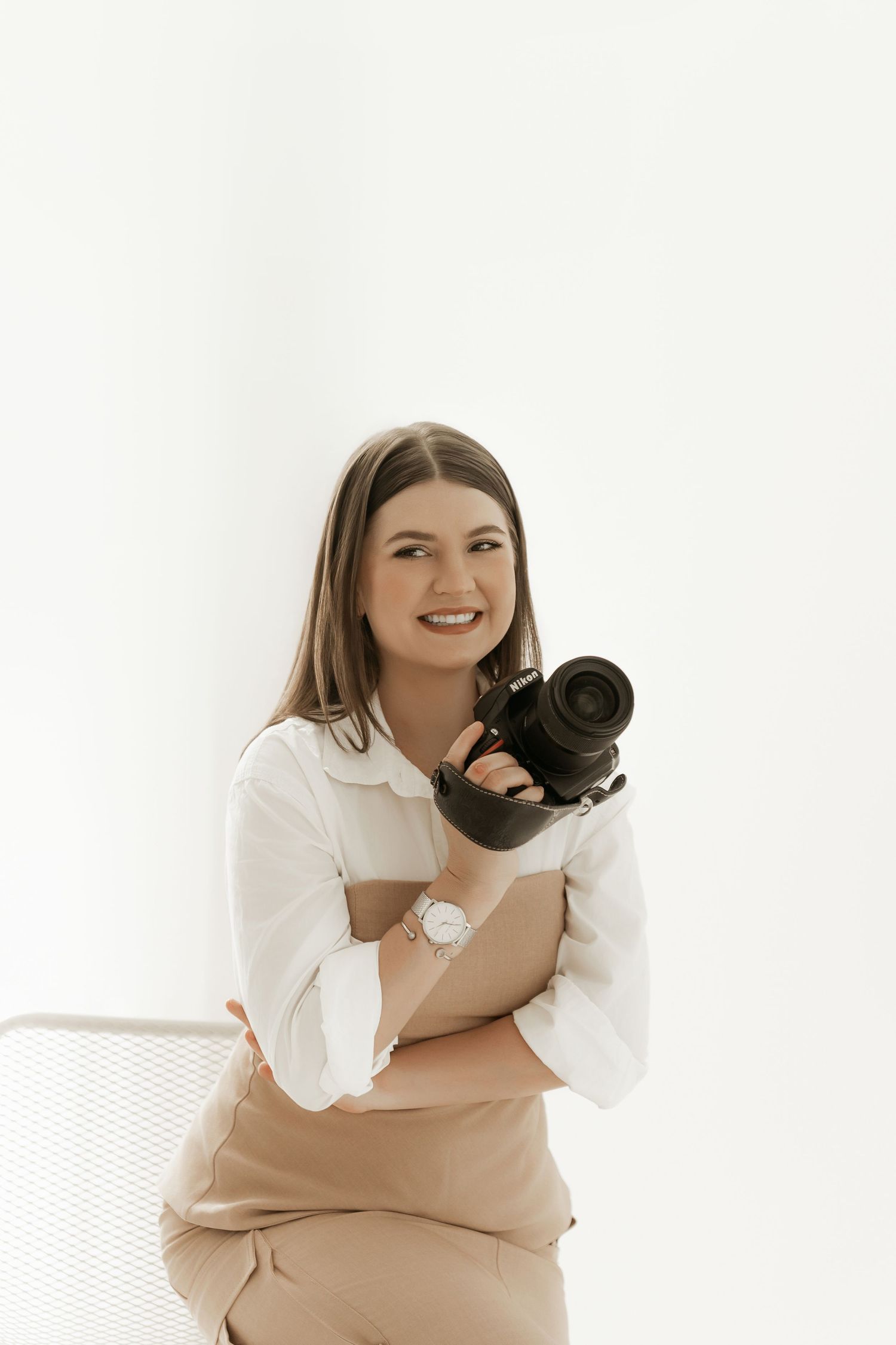 Professional photographer in white top and beige pants holds camera while posing against white background in studio.