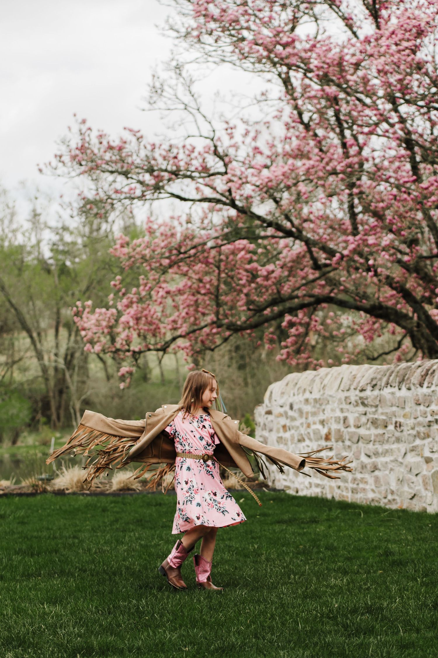 A series of three photos showing a child dancing and twirling under blooming pink cherry blossom trees by a stone wall.