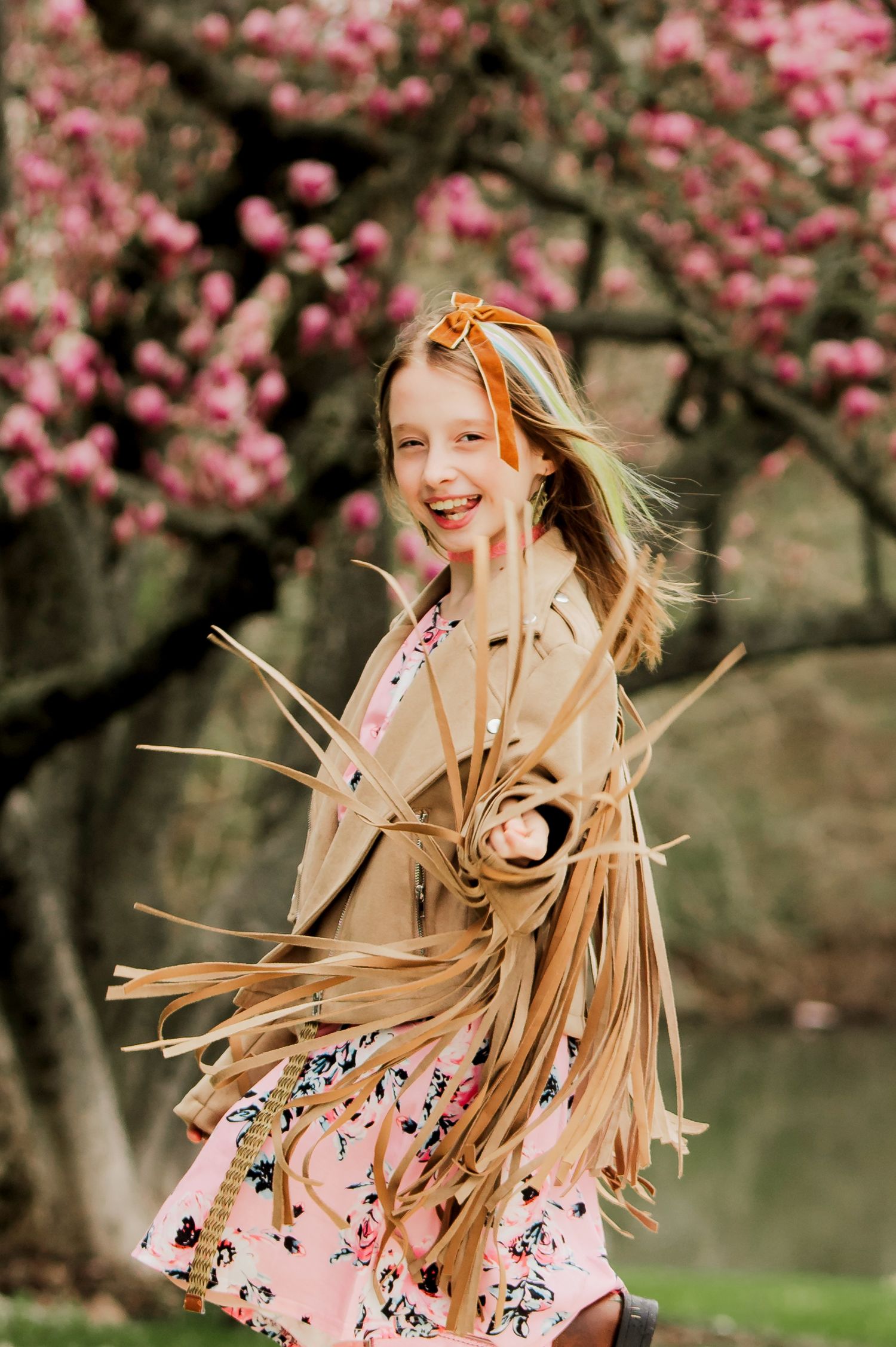 A person in a beige coat and pink dress spins joyfully beneath blooming cherry blossom trees in spring.
