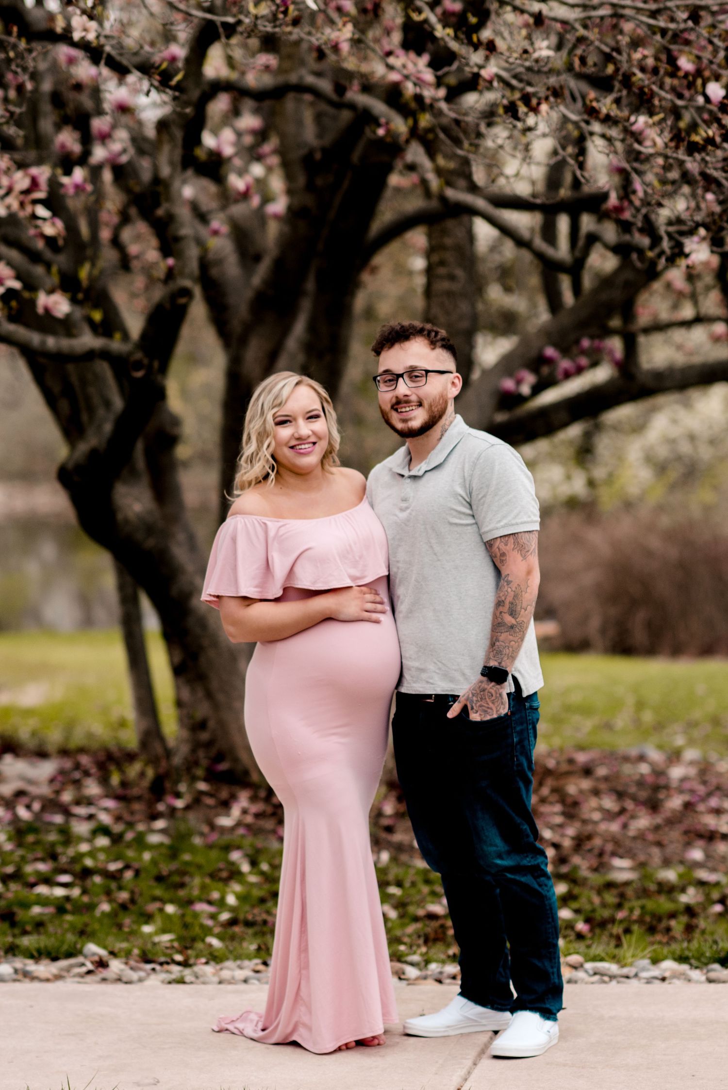 A couple poses under blooming cherry blossom trees, with the person in a pink dress showing a baby bump.