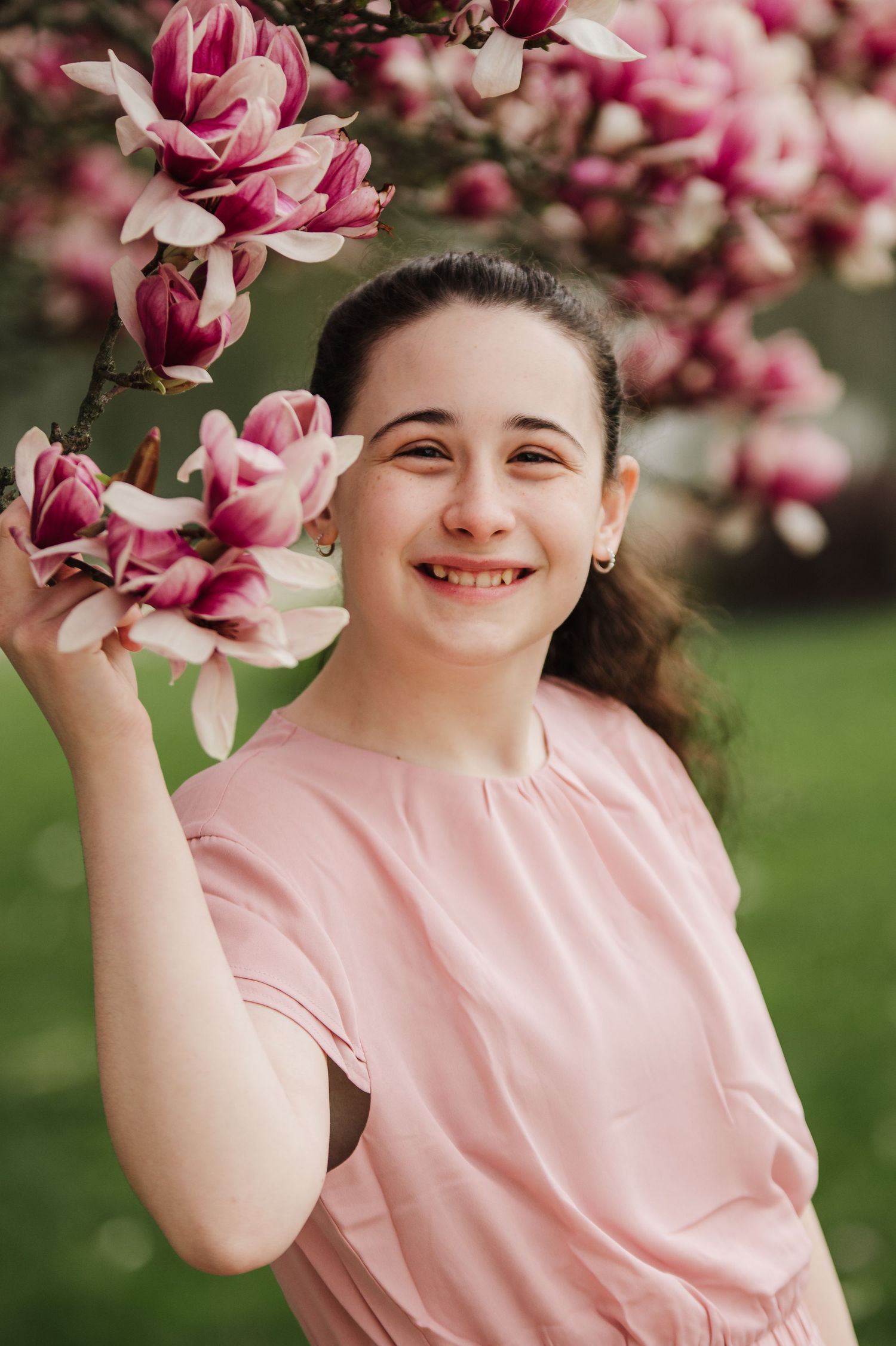 A person in a pink shirt smiles beneath blooming pink magnolia blossoms in a spring garden setting.